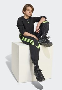 Black sweatshirt with green logo, black track pants with neon green stripes, black sneakers, and coordinating black socks. Seated on a cube.