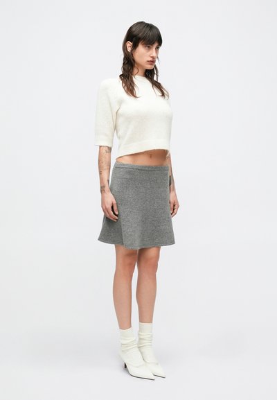 Λευκό κοντό πουλόβερ, γκρι φούστα με καμπάνα και λευκά ανδρικά μποτάκια με τακούνι. Απαλές υφές με ορατή ραφή. Το μοντέλο στέκεται μπροστά σε απλό φόντο.