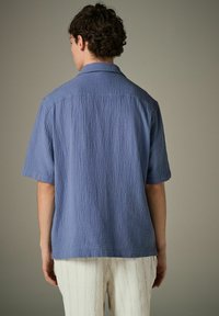 Kurzarmhemd aus strukturiertem hellblauem Stoff, mit klassischem Kragen und geradem Saum, von hinten gezeigt.