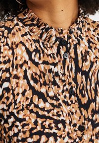 Brun och svart mönstrad blus med volangkrage, med fyra svarta knappar längs knäppslån och ett strukturerat tyg.