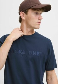 Jūras zila t-kreklu ar īsām piedurknēm ar iepresētu tekstu "JACK & JONES PREMIUM". Gluda kokvilnas auduma. Brūna cepure nēsāta nedaudz slīpi.