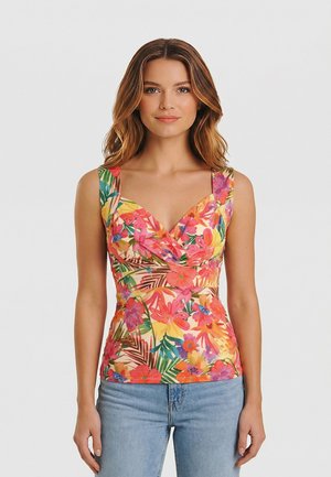 Giovane donna che indossa un top senza maniche floreale colorato con scollo a V e jeans azzurri, in piedi davanti a uno sfondo chiaro uniforme.