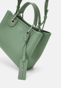 Zielona torba typu tote z ziarnistej skóry z podwójnymi rączkami, srebrnymi okuciami i skórzaną metką z logo. Charakteryzuje się usztywnionym kształtem i fakturowaną powierzchnią.