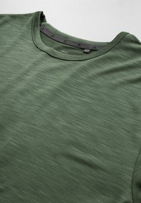 Gros plan sur un T-shirt vert à col rond avec une texture subtile et un détail de biais intérieur noir au niveau du col.