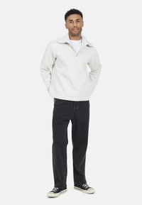 Lichtgrijze ritsloze pullover van zachte stof, met een ontspannen pasvorm en hoge kraag, gecombineerd met zwarte spijkerbroek en zwarte sneakers.