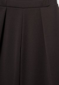 Mørkebrun skræddersyet stof med strukturerede syninger og folder, sandsynligvis en del af et tøjstykke såsom bukser eller et skørt.