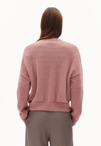 Eine Person mit langen braunen Haaren trägt einen rosa Strickpullover und graue Hosen und steht mit dem Rücken zu einem schlichten weißen Hintergrund.
