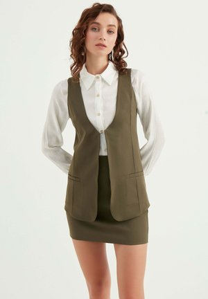 Olijfgroene, gestructureerde vest met een diepe V-hals en twee voorzakken, gelaagd over een witte blouse met kraag, afgewerkt met een getailleerde rok.