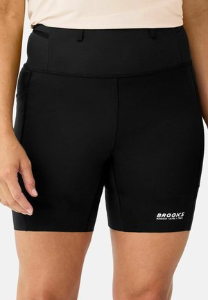 Nahaufnahme einer Person, die schwarze Brooks-Laufshorts mit hohem Bund und Logo am linken Oberschenkel trägt.