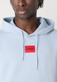 Svetlo modrá mikina s kapucňou s jemnou textúrou a predným červeným obdĺžnikovým logom, na ktorom je nápis "HUGO". Obsahuje šnúrky na kapucni.