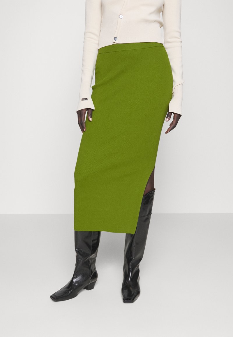 Mannequin portant une jupe midi verte avec une fente latérale, un haut crème à manches longues boutonné et des bottes noires hautes jusqu'aux genoux.