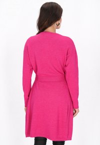 Nő élénk rózsaszín kardigánt visel, övvel, háttal áll, hosszú ujjai és fülbevalói látszanak.