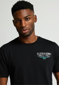 Camiseta de algodón negra con bordado blanco y turquesa que incluye "ALESSANDRO Milano Italia" y detalles de ubicación. Cuello redondo, mangas cortas.