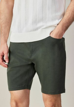 Mann trägt dunkelgrüne knielange Shorts und ein weißes, gestricktes ärmelloses Oberteil, mit einer Hand in der Tasche und der anderen entspannt an der Seite.