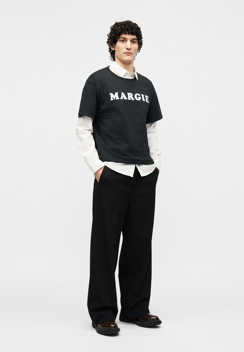 Jeune homme aux cheveux bouclés portant un t-shirt noir Margiel superposé à une chemise blanche, un pantalon noir à jambes larges et des chaussures marron, debout les mains dans les poches.