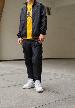 Personne portant un survêtement noir avec des baskets blanches et un t-shirt jaune avec un logo de basket-ball noir, debout sur un trottoir gris.