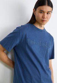 Μπλε βαμβακερό t-shirt με υφαντό λογότυπο Timberland και έμβλημα δέντρου μπροστά. Διαθέτει κοντά μανίκια και στρογγυλή λαιμόκοψη.