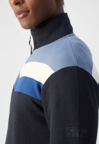 Pulover cu fermoar la jumătate, cu un design în blocuri de culoare negru, albastru și crem. Fabricat dintr-un material texturat, cu text brodat pe mânecă.