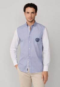 Uomo con capelli ricci che indossa una camicia a righe blu e bianche con maniche bianche e pantaloni beige, in piedi contro uno sfondo semplice.