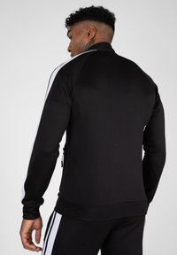 Zwarte sportieve jas met een hoge kraag, witte zijstrepen, ritszakken en aansluitende manchetten. Gestructureerde stof met een gladde afwerking.