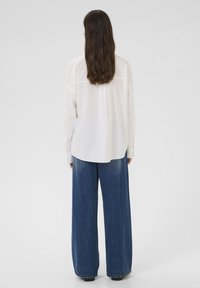 White long-sleeve shirt with a pleat detail at the back, paired with wide-leg blue denim jeans and black shoes.
