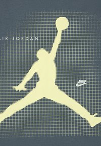 Σκούρο μπλε βαμβακερό t-shirt με κίτρινη ψηφιακή σιλουέτα ενός ατόμου που πηδάει, μοτίβο πλέγματος και το κείμενο "AIR JORDAN" με μικρό λογότυπο της Nike.