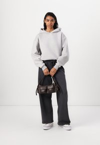 Felpa grigia oversize, pantaloni della tuta neri e sneakers bianche. Tiene in mano una piccola borsa in pelle marrone scuro con due tasche frontali.