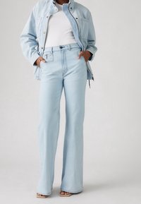 Person wearing light blue denim jacket and high-waisted wide-leg jeans over a white ribbed top, with hands in pockets and beige sandals.