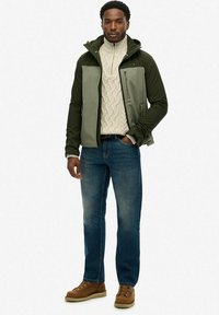 Olijfgroen zip-up jack over een crèmekleurige kabel gebreide trui, gecombineerd met blauwe jeans en bruine veterschoenen. Eenvoudig ontwerp, gestructureerde stoffen.