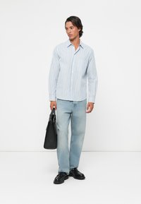 Man wearing white and blue striped shirt, light blue jeans, black shoes, holding black leather bag, standing against white background.