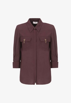 Blusa bordeaux con colletto, due tasche sul petto e dettagli in metallo dorato. Maniche lunghe ribaltate fino ai gomiti, tessuto morbido con una texture liscia.