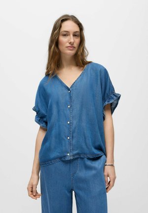 Vrouw draagt een loszittend denim overhemd met korte mouwen en ruches aan de mouwen, gecombineerd met bijpassende denim broek, staand tegen een effen achtergrond.