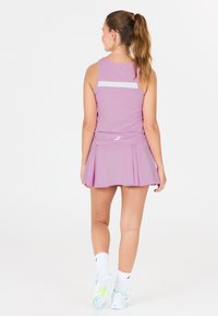 Giovane donna in canotta sportiva rosa e gonna a pieghe che cammina, indossando calze bianche e sneaker, su sfondo bianco uniforme.