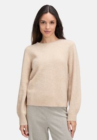 Frau in einem beigen Rundhalsausschnitt-Pullover und karierten Hosen, die vor einem schlichten hellen Hintergrund steht, nach vorne schaut und leicht lächelt.