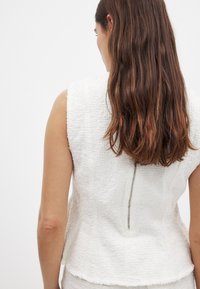 Hvid tekstureret ærmeløs top med lynlås bagpå. Stoffet ser blødt og taktilt ud, med et subtilt mønster nær skuldrene.