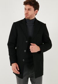 Melnas vilnas virsjakas ar iegrieztu apkakli, priekšējiem pogām un elegantu, pielāgotu piegriezumu. Stilizēta virs pelēka turtleneck un pelēkiem biksēm.