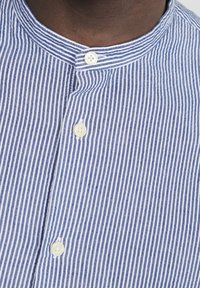 Sininen ja valkoinen raidallinen paita, jossa on aukoton kaulus, sisältäen kaksi valkoista nappia ja pehme�än pinnan. Raidat ovat tasaisesti seurattuja pystysuunnassa.