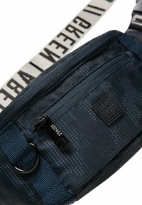 Navyblaue Umhängetasche aus strapazierfähigem Material, mit einem strukturierten Gittermuster, einer Reißverschlusstasche und einem kontrastierenden Schultergurt mit Textelementen.