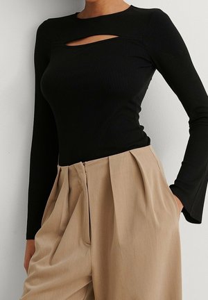 Vrouw draagt zwarte top met lange mouwen en uitsnijding op de borst, beige plissébroek met hoge taille, handen in de zakken, tegen een effen achtergrond.