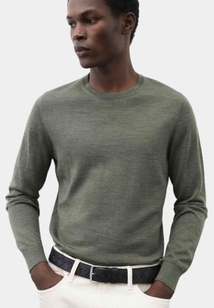 Man wearing an olive green long-sleeve sweater tucked into white pants with a black belt, hands in pockets, looking to the side.