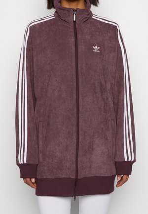 Person iført en bordeaux Adidas træningsjakke med hvide striber på ærmerne og lynlås foran, kombineret med hvide bukser.