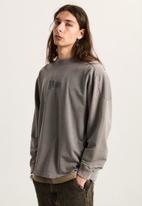 Šedé tričko z bavlny s dlouhým rukávem, kulatým výstřihem a vyšitým designem na hrudi, doplněné o tmavé kalhoty pro kontrast.