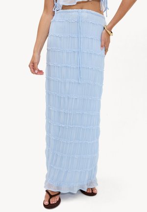 Femme portant une jupe longue bleu clair texturée avec une ceinture élastique à nouer, des sandales marron, un bracelet doré et un haut court blanc.