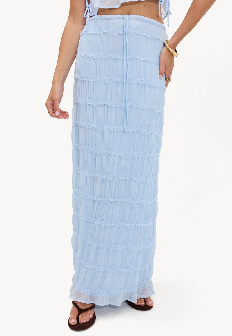 Femme portant une jupe longue bleu clair texturée avec une ceinture élastique à nouer, des sandales marron, un bracelet doré et un haut court blanc.