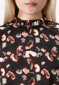 Blouse à motifs sombres avec un col volanté. Présente un mélange de formes abstraites brun, noir et crème sur un fond bordeaux.