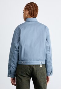Giacca cropped azzurra realizzata in tessuto resistente. Presenta accessori in metallo argentato e un colletto, abbinata a pantaloni cargo a motivo verde scuro.