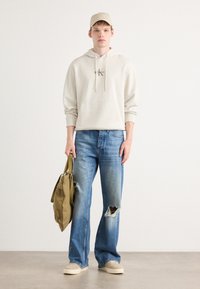 Beige Kapuzenpullover mit einer Vordertasche, ausgebleichte hellblaue Jeans, braune Sneakers und eine olivgrüne Tasche. Modell trägt eine passende Mütze.