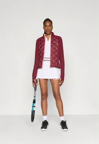 Burgundy athletic jacket with textured front, paired with a white dress featuring pink accents, black sneakers, and holding a tennis racket.