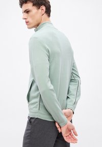 Ανοιχτό πράσινο, υφασμάτινο μπουφάν με φερμουάρ και ψηλό γιακά, με αντίθετες ραφές στα πλάγια. Φοράει γκρι παντελόνια, το μοντέλο ποζάρει με τα χέρια πίσω από την πλάτη.