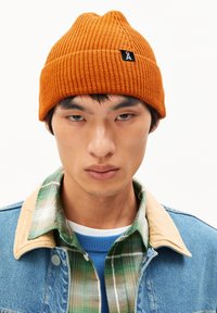 Orange knitted beanie with a textured ribbed pattern and a small black logo tag. Worn over a denim jacket with a plaid shirt collar.